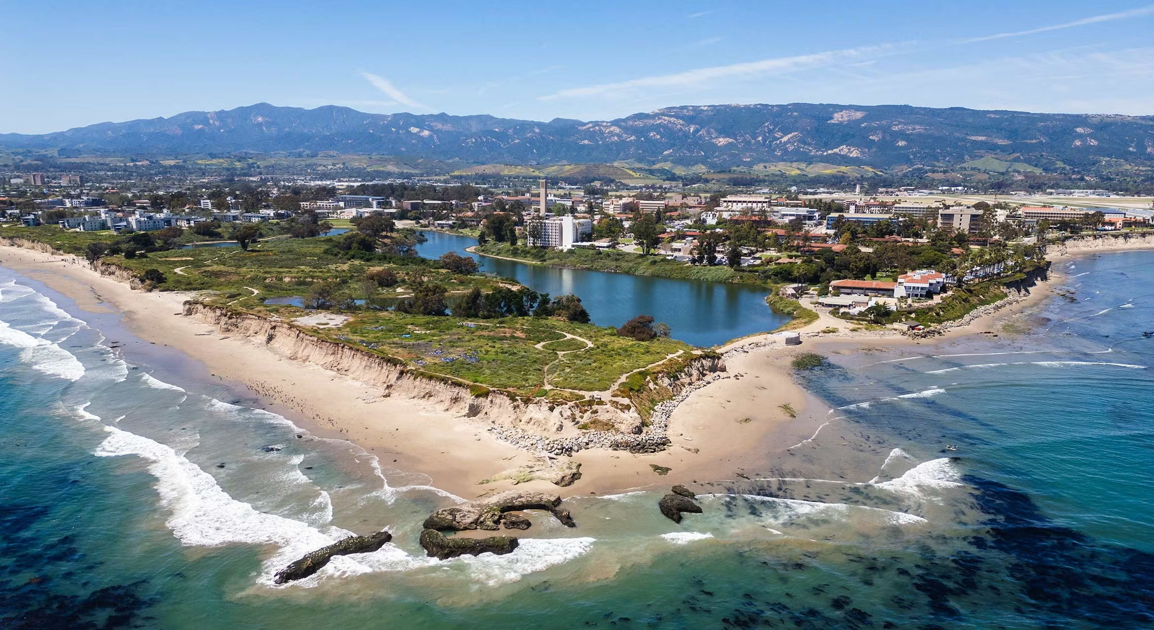 Aerial view of Campus Point at UC Santa Barbara, showing sandy cliffs and the beach meeting turquoise Pacific Ocean waters, with the campus lagoon, university buildings, and the Santa Ynez Mountains visible in the background under a clear blue sky.