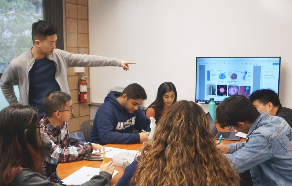 A group of students are gathered around a table working on materials during a small-group biology session. An instructor points toward a monitor in the background displaying biological images and diagrams.