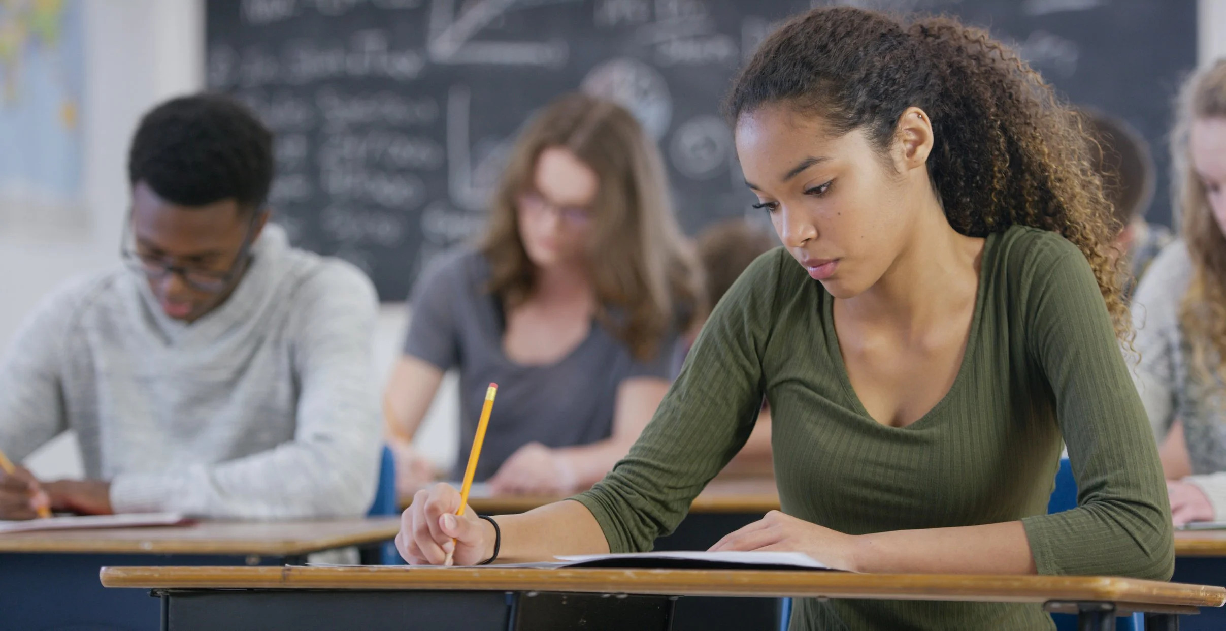 A focused student writing with a pencil at a classroom desk, with other students working in the background in front of a chalkboard.